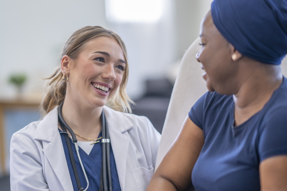 A female Oncologist meets with her cancer patent in the comfort of her own home as she checks up on her between treatments. She is wearing a white lab coat and holding out a clipboard as the two talk. The patient is seated in a high-back chair and wearing a headscarf to keep warm.